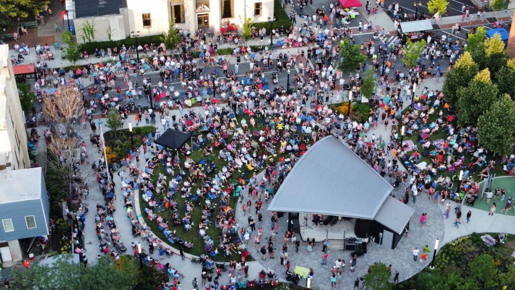 Image of a concert at a park, showing a large crowd gathering on the green space and under the trees